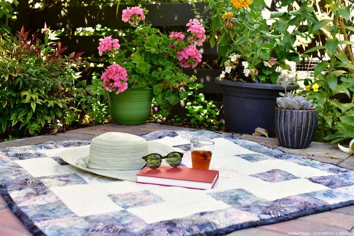 Picknickdecke im blühenden Garten mit Buch, Sonnenhut und Getränk, entspannte Sommeratmosphäre für eine gemütliche Staycation.