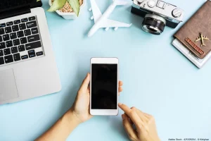 Eine Person hält ein Smartphone neben Laptop, Kamera, Flugzeugmodell und Reisepass, passend zum Thema Reisen planen App.